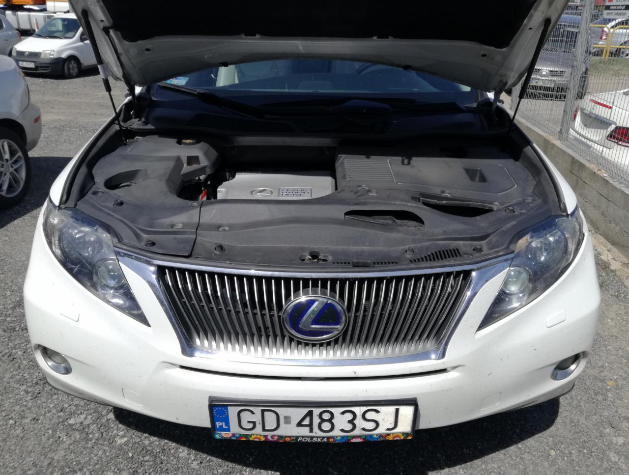 Biały Lexus z otwartą maską prezentuje silnik hybrydowy. Widoczny numer rejestracyjny. Ujęcie z przodu, na zewnątrz, w słoneczny dzień.