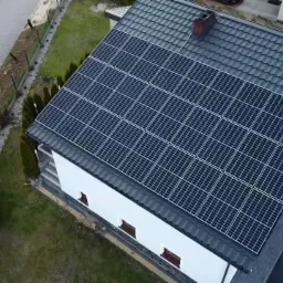 Widok z drona na dom z panelami fotowoltaicznymi na dachu z szarej dachówki, komin z cegły, biała elewacja, zadbany trawnik i podjazd z kostki brukowej.