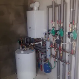 Kompletna instalacja centralnego ogrzewania z białym zasobnikiem ciepłej wody, kotłem gazowym i pionowymi rurami z zielonymi pompami obiegowymi, zamontowana na tle surowej ściany.