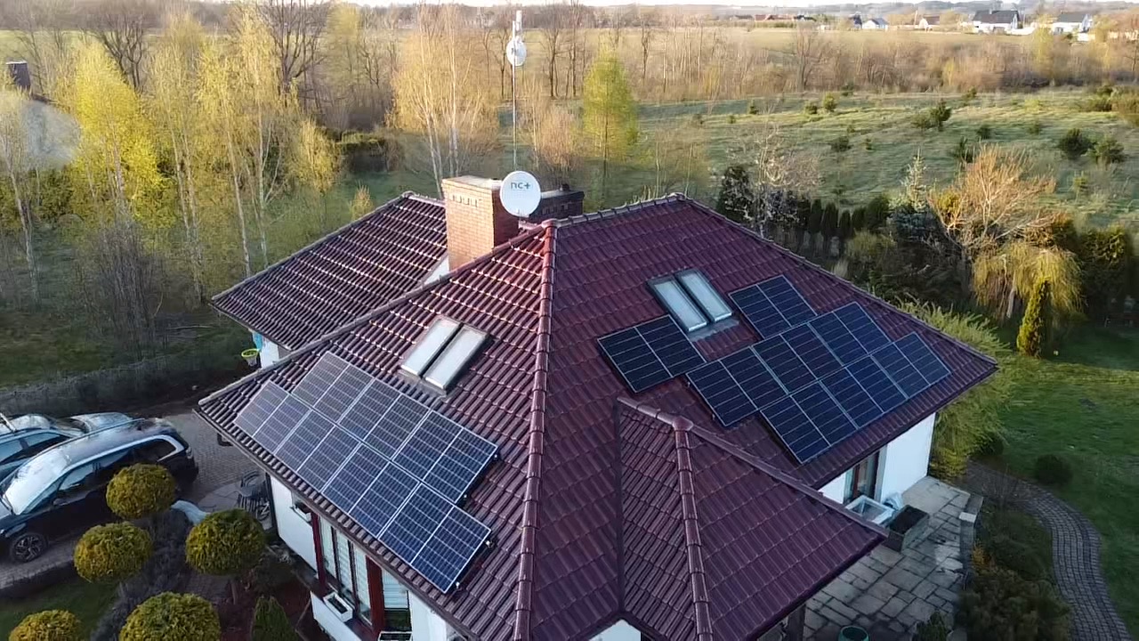 Widok z lotu ptaka na dom z czerwoną dachówką i panelami słonecznymi, antena satelitarna na kominie, w tle drzewa i pole.