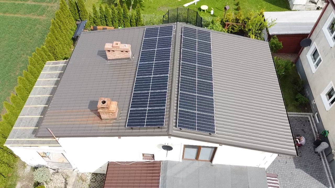 Widok z góry na dom z brązowym dachem, na którym zamontowane są dwa rzędy paneli fotowoltaicznych, obok komina z czerwonej cegły. W tle widoczny zielony ogród.