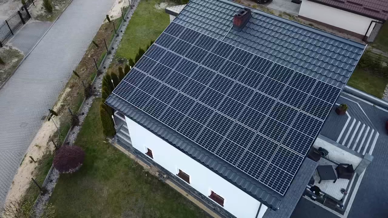 Widok z drona na dom z panelami fotowoltaicznymi na dachu z szarej dachówki, komin z cegły, biała elewacja, zadbany trawnik i podjazd z kostki brukowej.