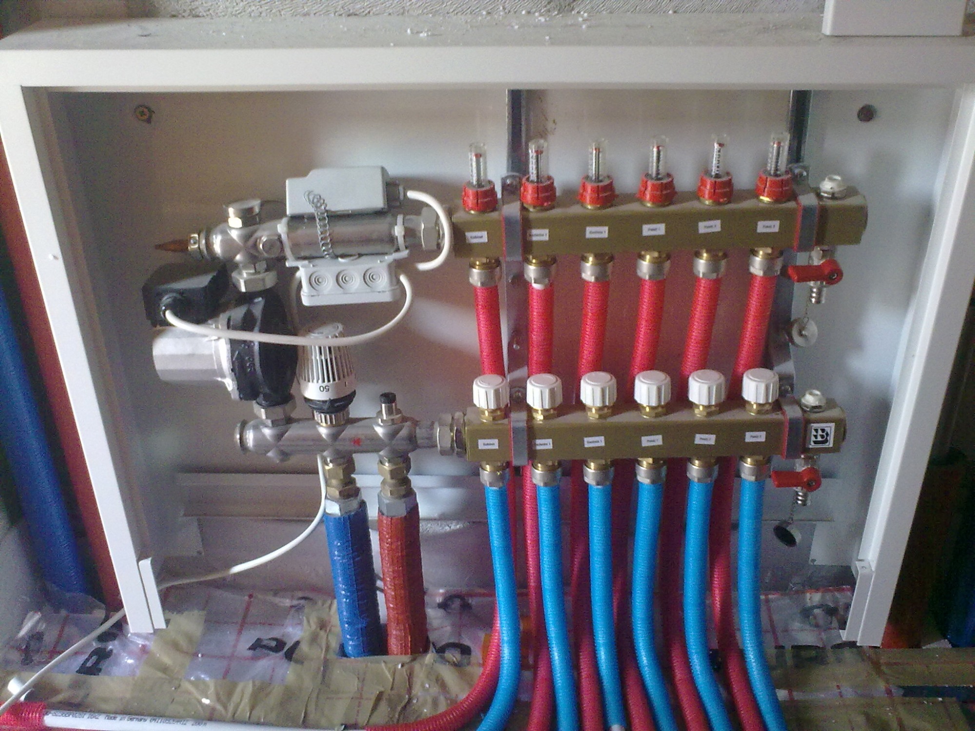 Rozdzielacz ogrzewania podłogowego w szafce, z widocznymi rurami w kolorach czerwonym i niebieskim, podłączonymi do listwy z zaworami i przepływomierzami.