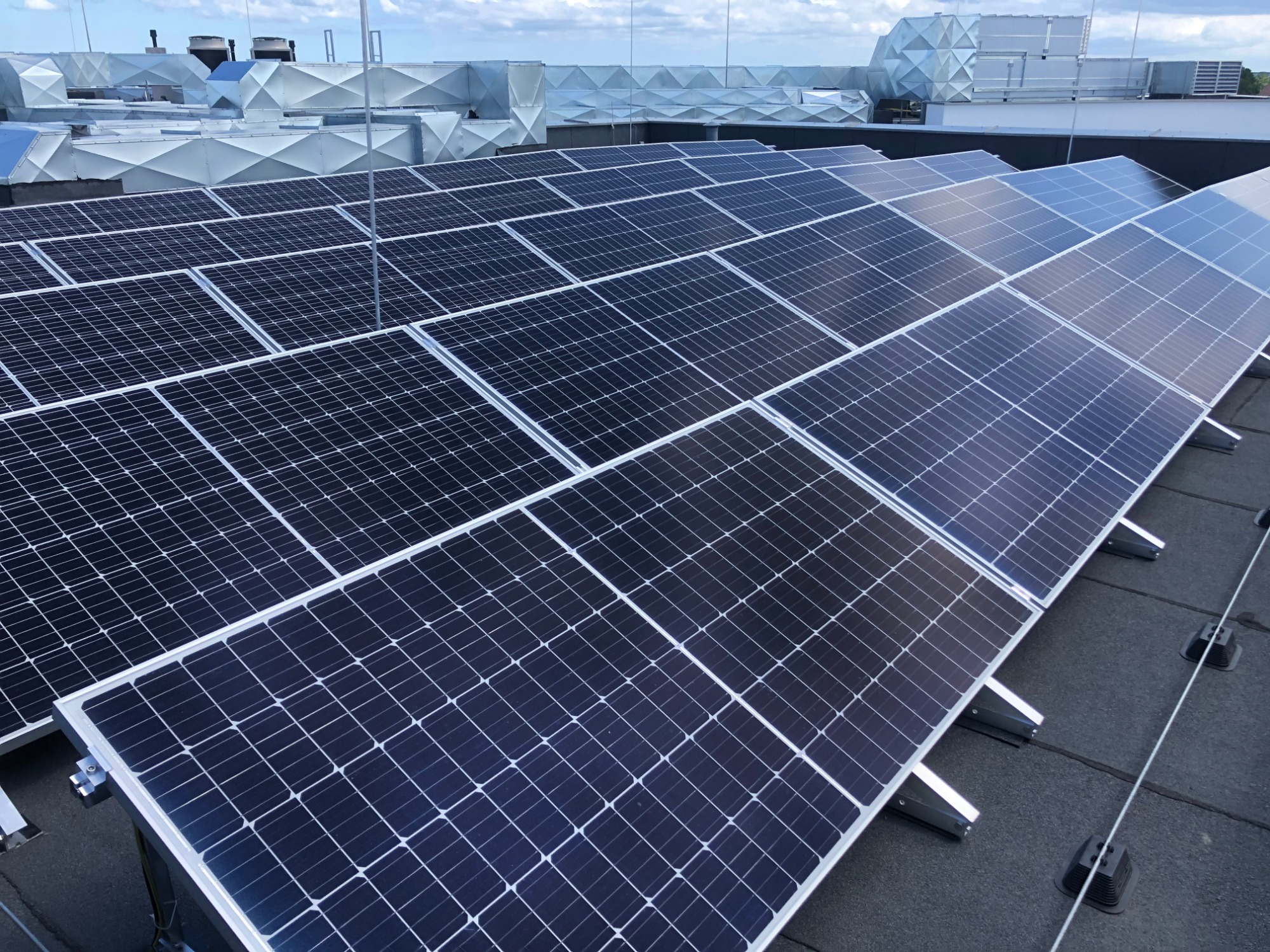 Rząd paneli fotowoltaicznych na płaskim dachu z widocznymi elementami wentylacji przemysłowej w tle, oświetlonych naturalnym światłem dziennym.