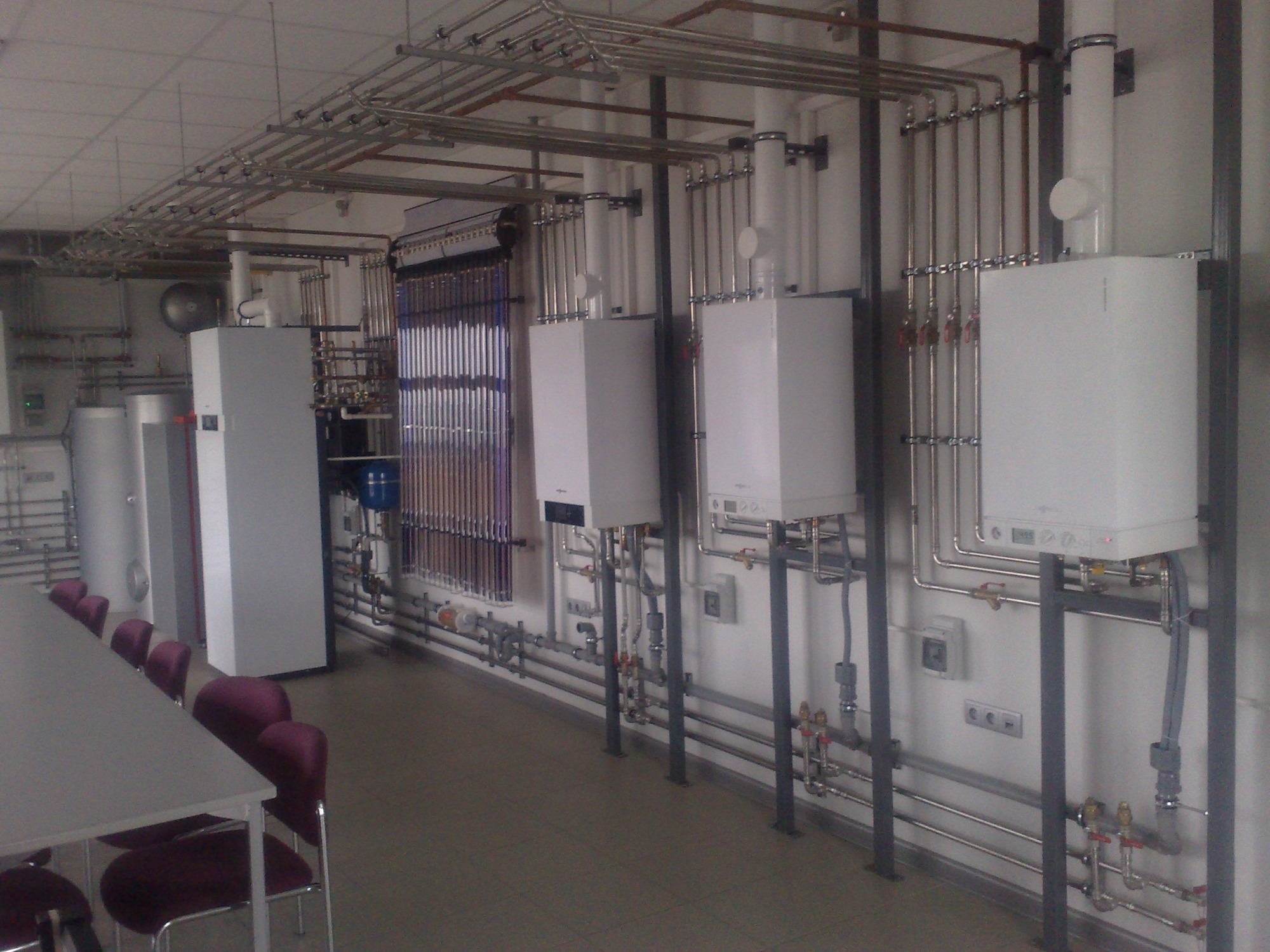 Instalacja grzewcza w pomieszczeniu biurowym, widoczne kotły grzewcze, rury, kolektory słoneczne i stół konferencyjny z krzesłami.