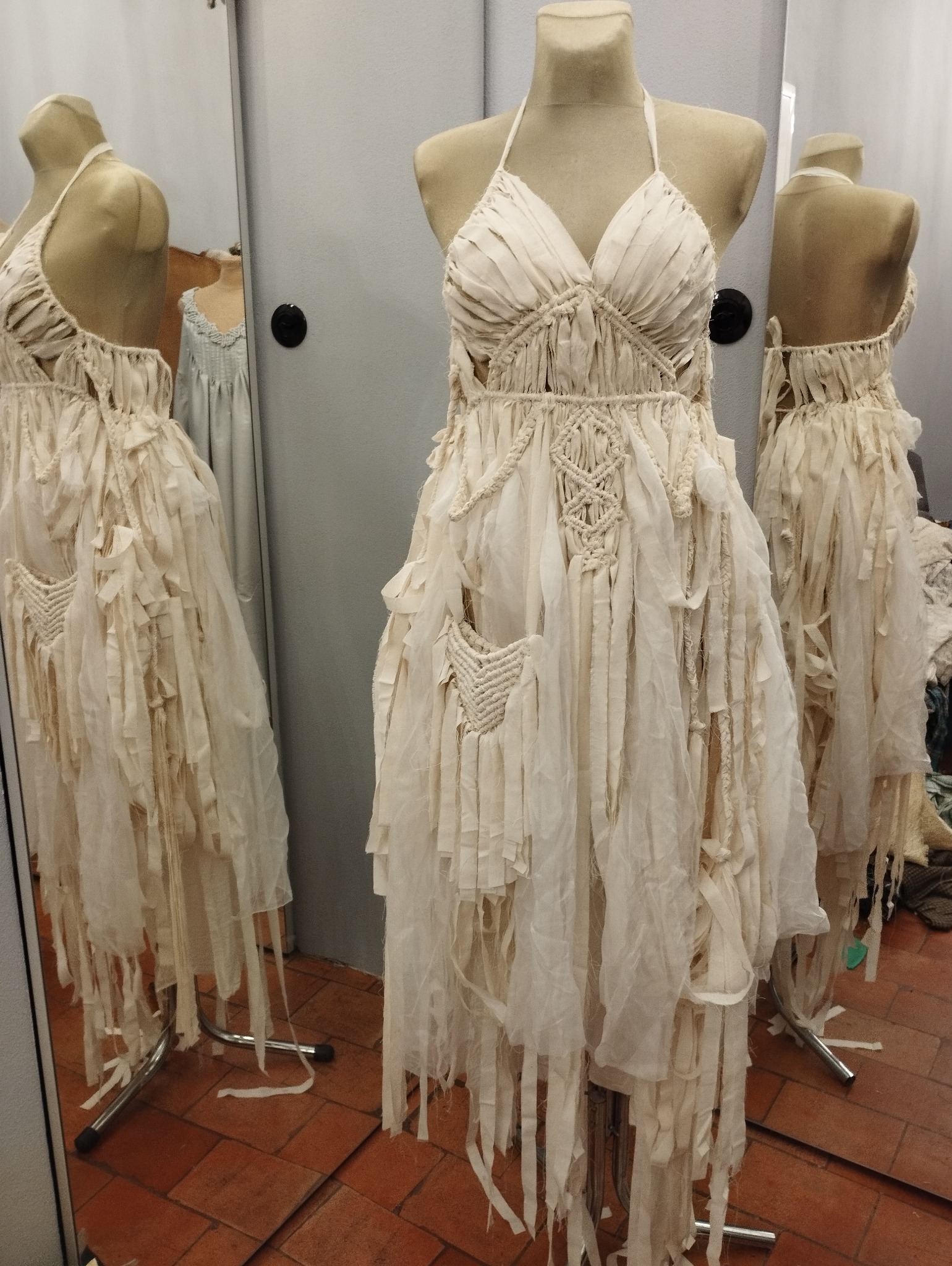 Kremowa suknia z wiszącymi paskami materiału i elementami makramy, prezentowana na manekinie w szwalni z lustrami.