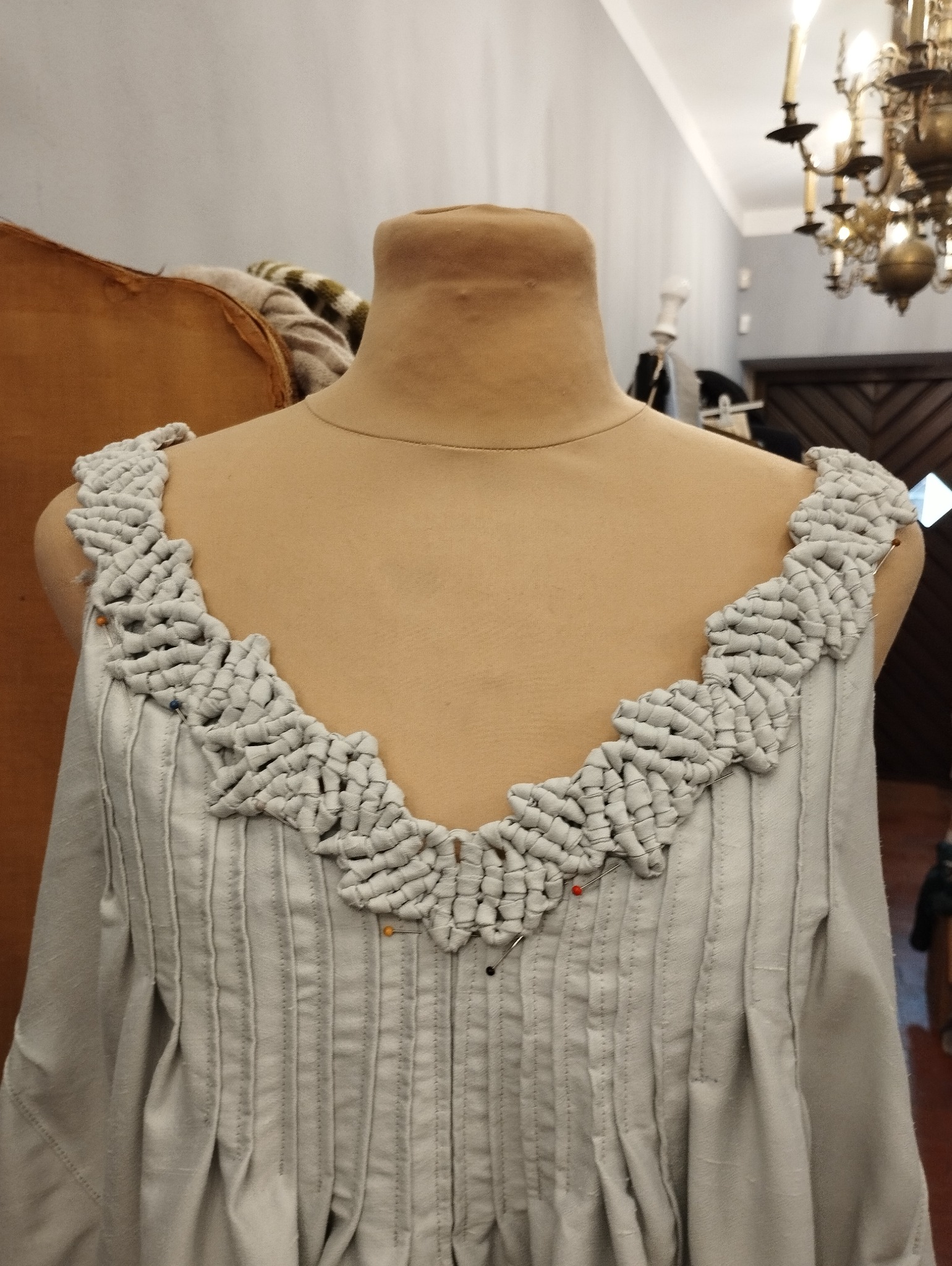 Biała suknia z dekoracyjnym, plecionym dekoltem na manekinie krawieckim, widoczne szpilki, w tle żyrandol.