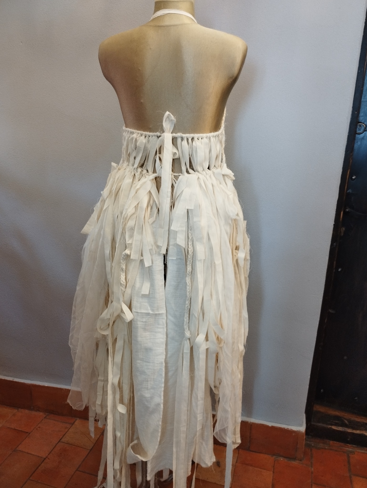 Sukienka z postrzępionego, kremowego materiału na manekinie krawieckim, widok z tyłu, z detalami wiązania i dekoracyjnych pasków.