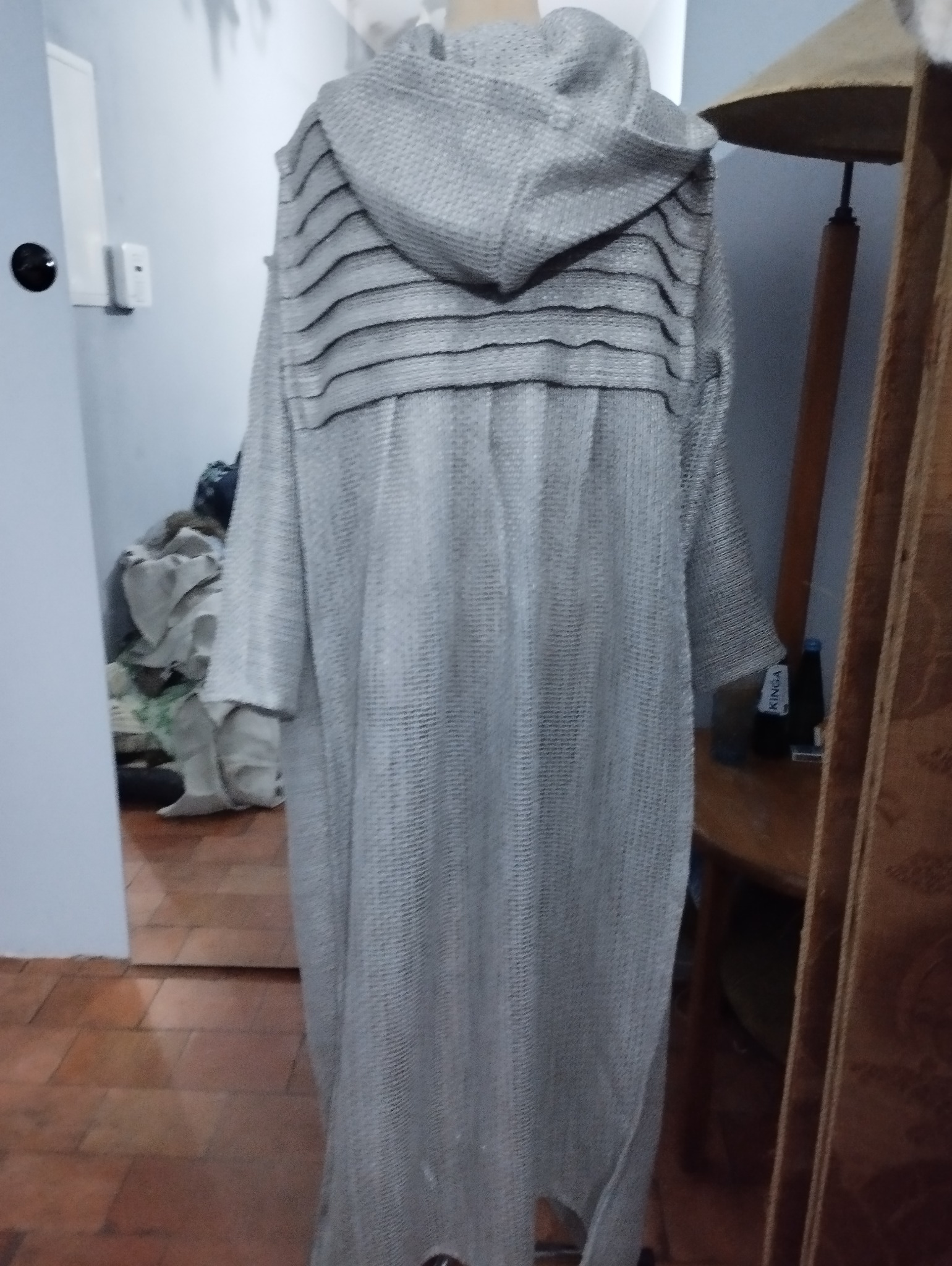 Długi, szary płaszcz z kapturem i poziomymi, czarnymi paskami na plecach, wiszący na manekinie w pomieszczeniu z terakotową podłogą.