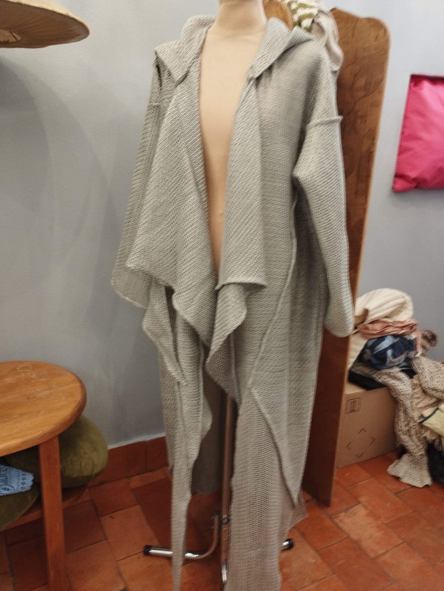 Długi, szary kardigan z kapturem, wykonany z dzianiny o wyraźnej fakturze, prezentowany na manekinie na tle ściany i elementów wyposażenia pracowni.