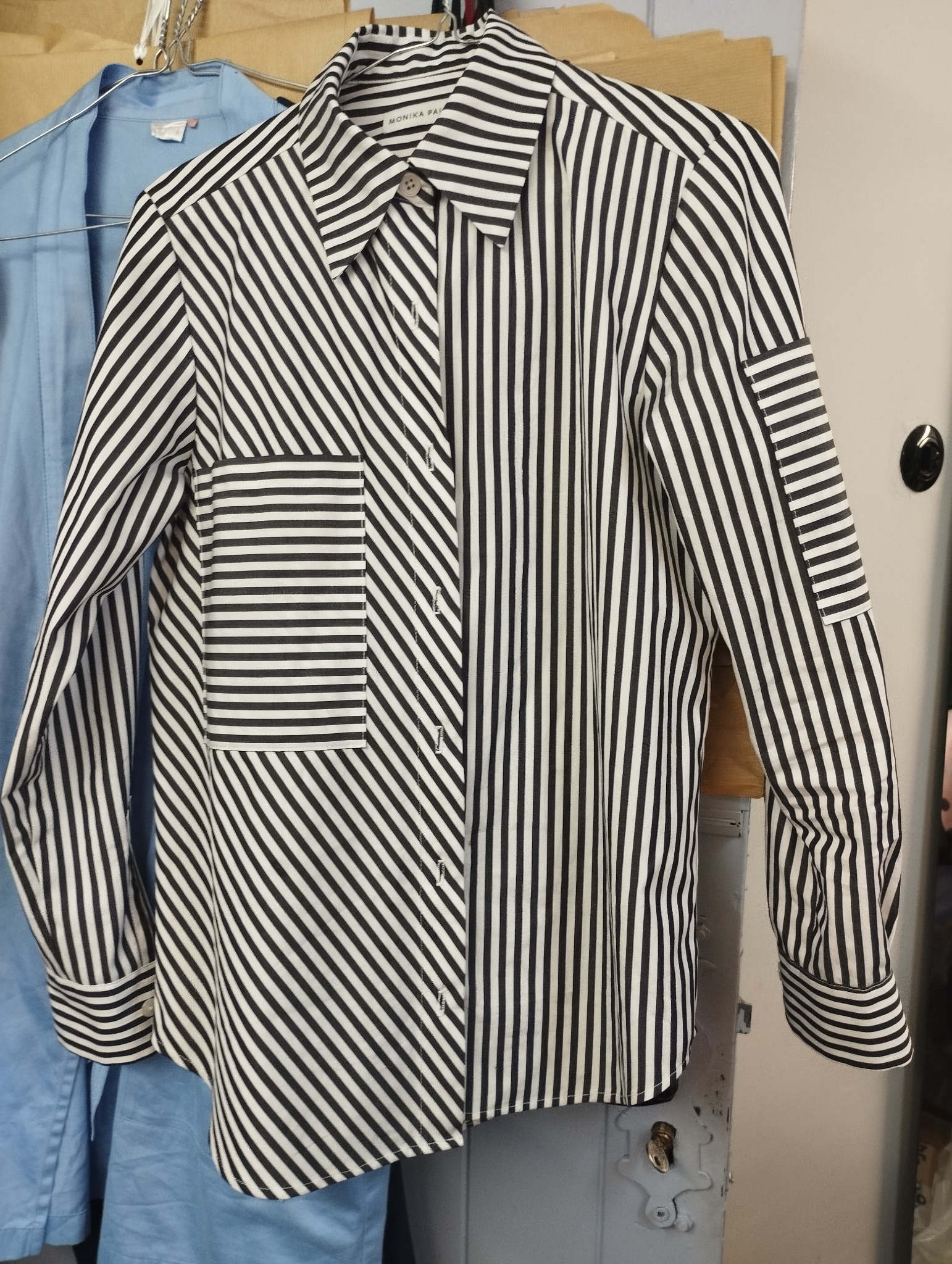 Czarno-biała koszula w paski z kieszenią na piersi, wisząca na wieszaku obok błękitnej koszuli.