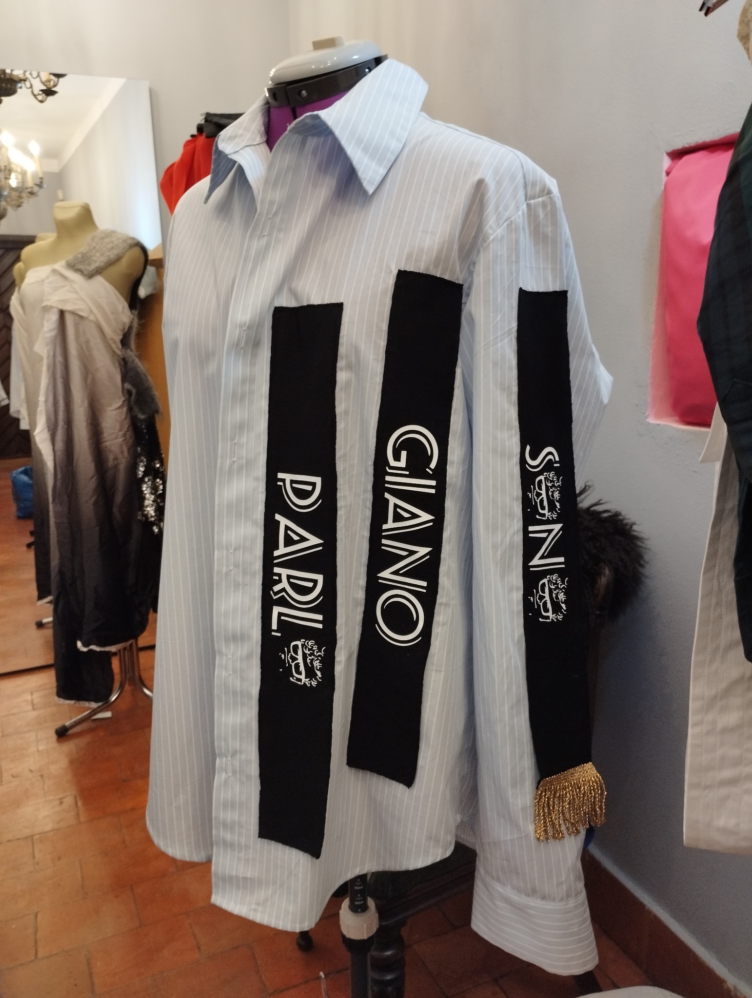 Koszula w paski z naszytymi czarnymi pasami z białymi napisami i złotymi frędzlami na manekinie krawieckim, widoczne inne ubrania w tle.