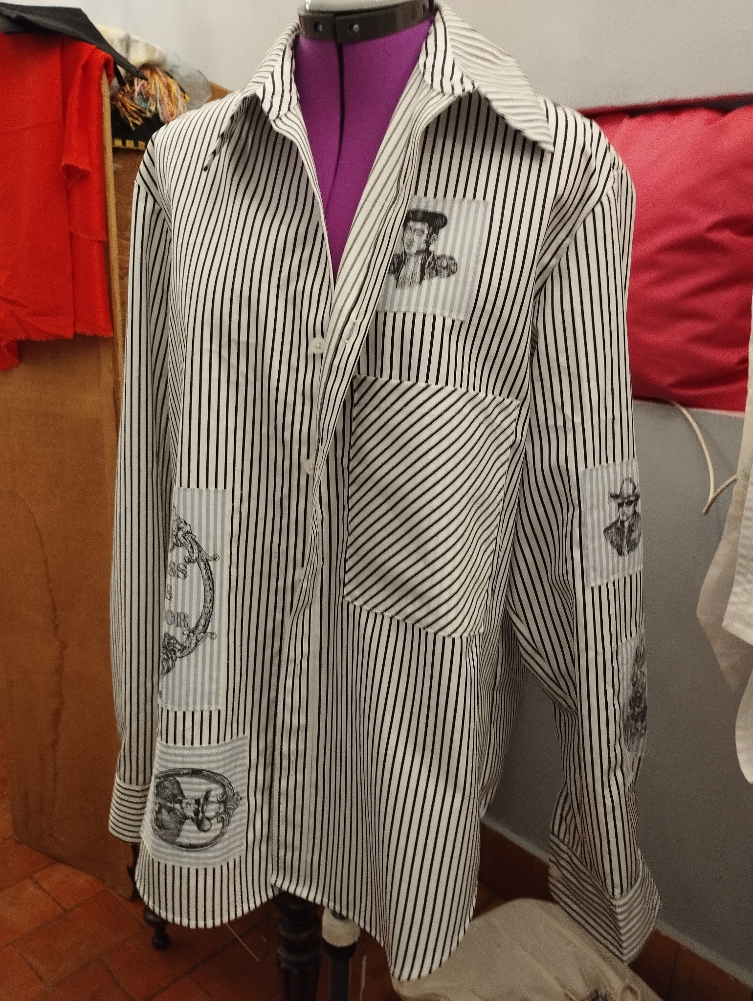 Czarno-biała koszula w paski z naszytymi grafikami na manekinie krawieckim, widoczne detale graficzne i nietypowe rozmieszczenie wzorów.