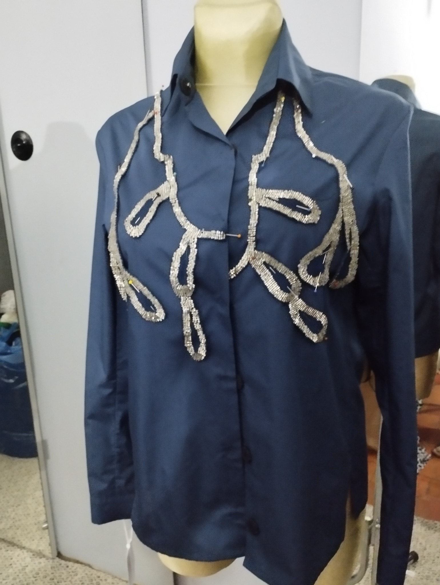 Granatowa koszula na manekinie z przypiętymi szpilkami srebrnymi elementami dekoracyjnymi układającymi się we wzór roślinny w trakcie tworzenia.