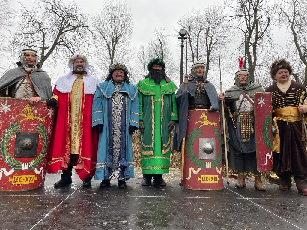 Grupa mężczyzn w kostiumach historycznych, z tarczami i włóczniami, na scenie podczas przedstawienia plenerowego.