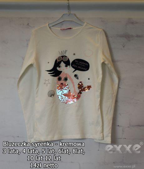 Kremowa bluzka z długim rękawem, ozdobiona motywem syrenki z cekinowym ogonem, zawieszona na wieszaku na tle ściany imitującej cegły.