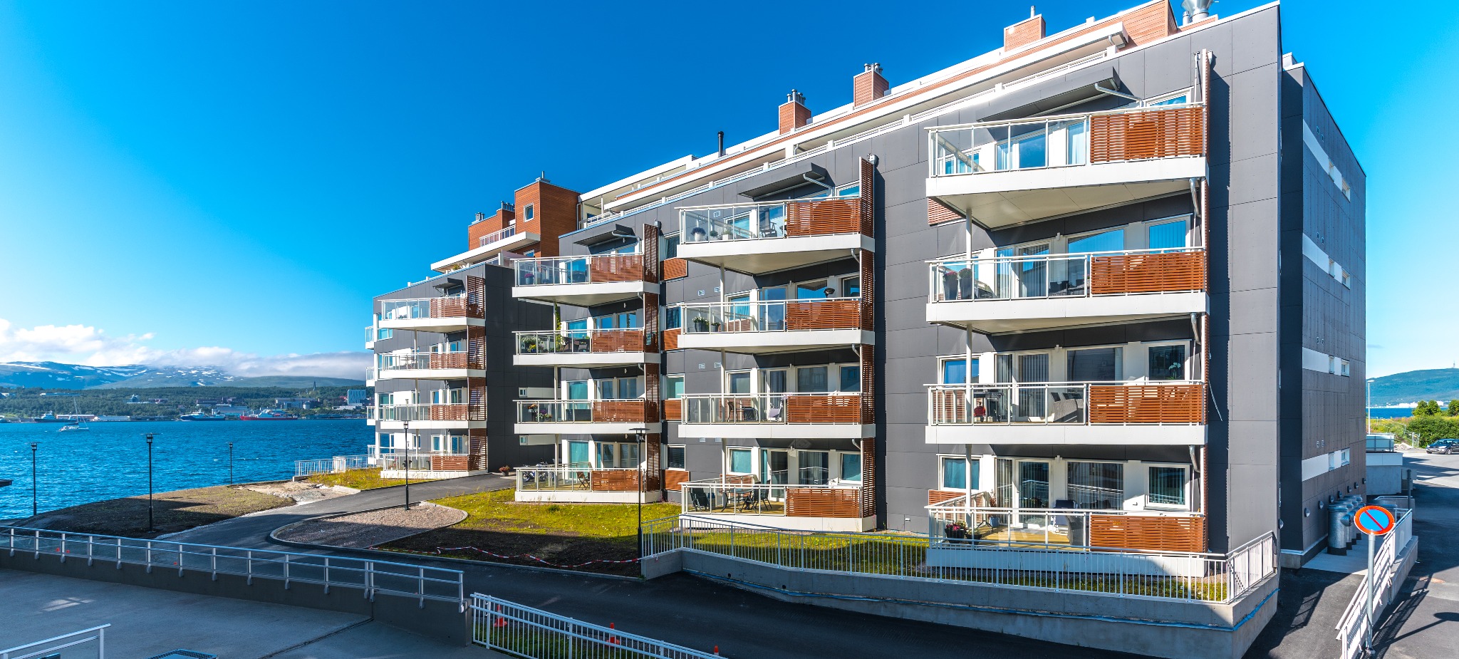 Nowoczesny, wielopiętrowy budynek mieszkalny z balkonami, szara elewacja, drewniane elementy, widok na morze i góry w tle, słoneczny dzień.