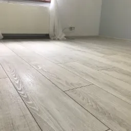 Jasne panele podłogowe z widoczną strukturą drewna, ułożone w nowym mieszkaniu, z grzejnikiem i zasłonami w tle.