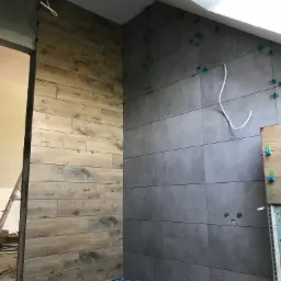 Montaż płytek: Ściana wyłożona szarymi płytkami i druga z drewnopodobnymi w trakcie układania, widoczne niebieskie krzyżyki dystansowe, wystające kable i otwory na gniazdka elektryczne.