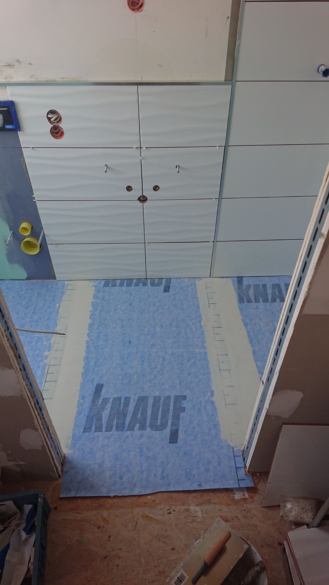 Białe płytki ścienne z falistym wzorem, częściowo ułożone w łazience, z widoczną folią hydroizolacyjną Knauf na podłodze i wystającymi rurami