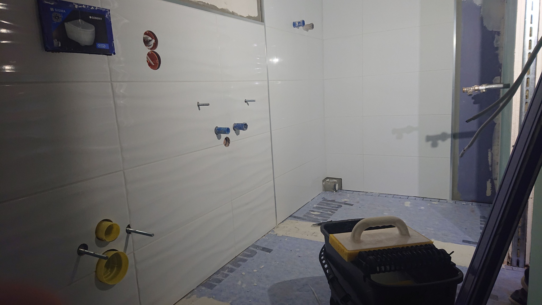 Wnętrze łazienki w trakcie prac wykończeniowych: białe płytki na ścianach, widoczne instalacje wodne, narzędzia glazurnicze w wiadrze, oraz opakowanie podtynkowego stelaża WC.