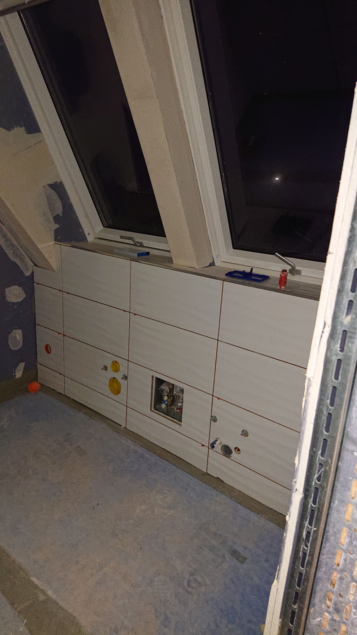 Ściana łazienki w trakcie kładzenia jasnych, kwadratowych płytek ceramicznych, widoczne otwory na instalacje, okno dachowe i narzędzia glazurnicze.