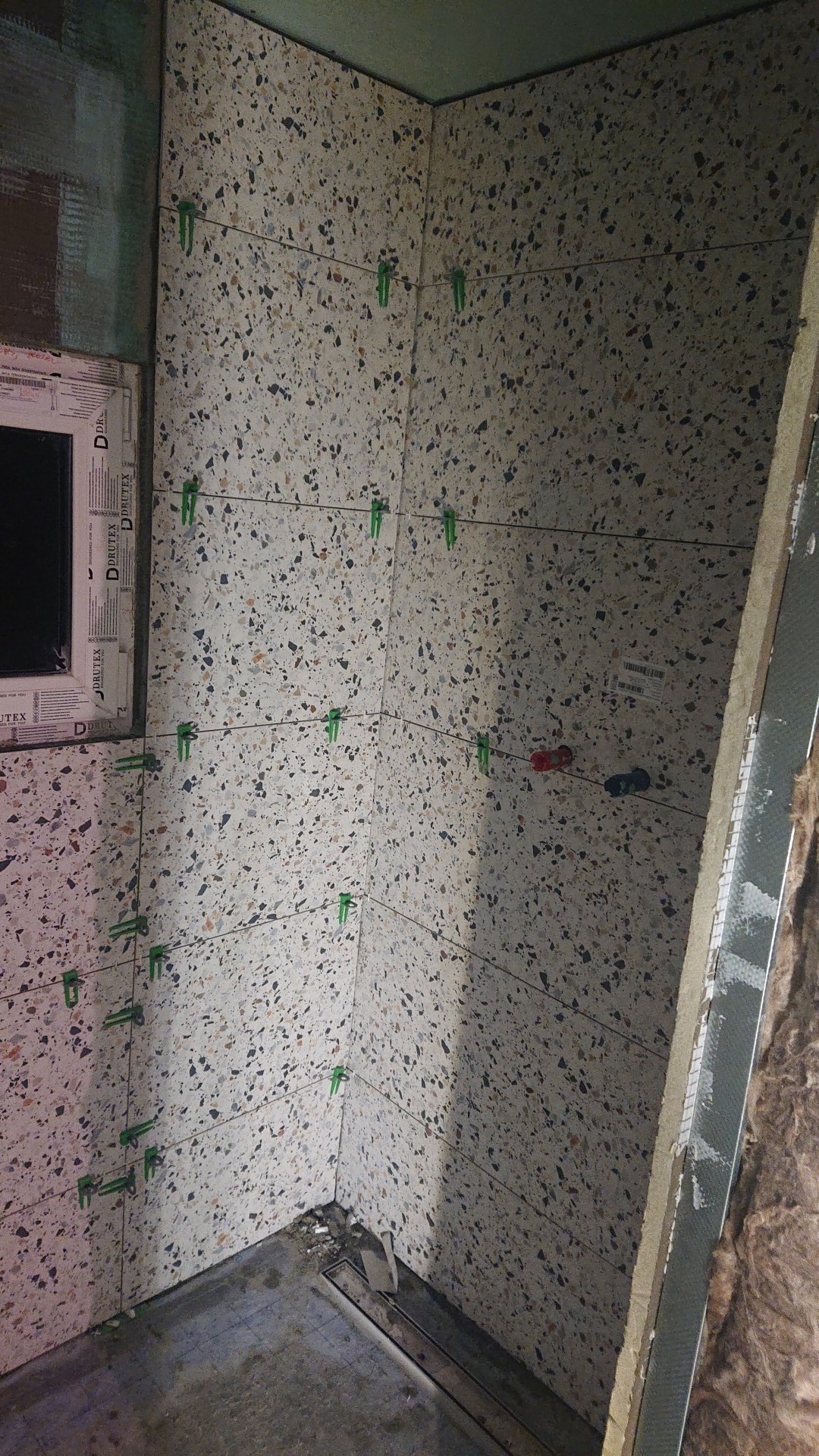 Ściany w trakcie wykańczania glazurą imitującą lastryko, z użyciem klipsów poziomujących, widoczny odpływ liniowy w podłodze i fragment ściany z oknem.