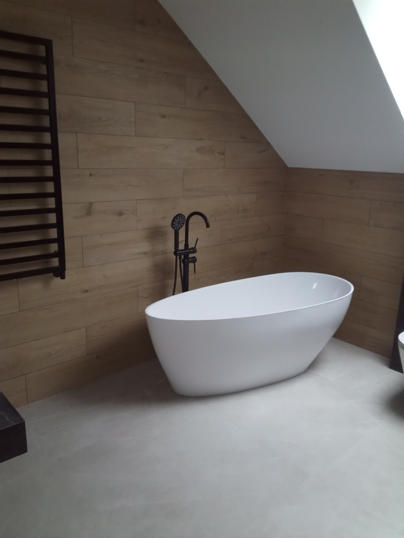 Nowoczesna łazienka na poddaszu z wolnostojącą białą wanną, czarną armaturą i grzejnikiem drabinkowym na ścianie wyłożonej panelami imitującymi drewno.