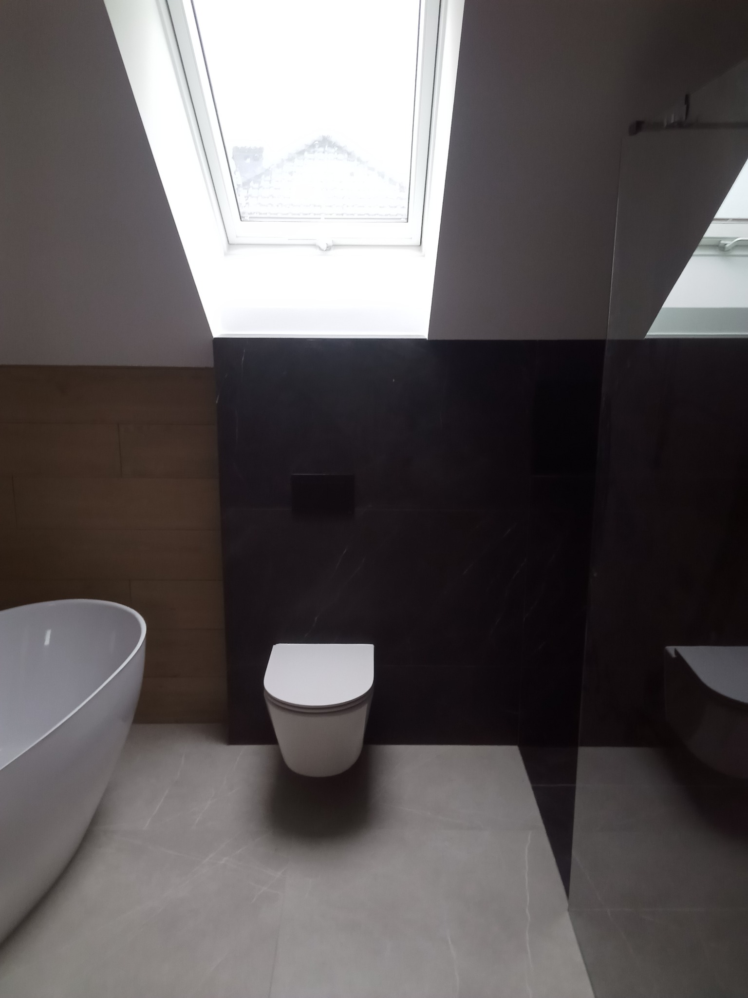 Nowoczesna łazienka na poddaszu z oknem dachowym, owalną wanną, wiszącą toaletą i czarnymi płytkami imitującymi marmur na ścianie.