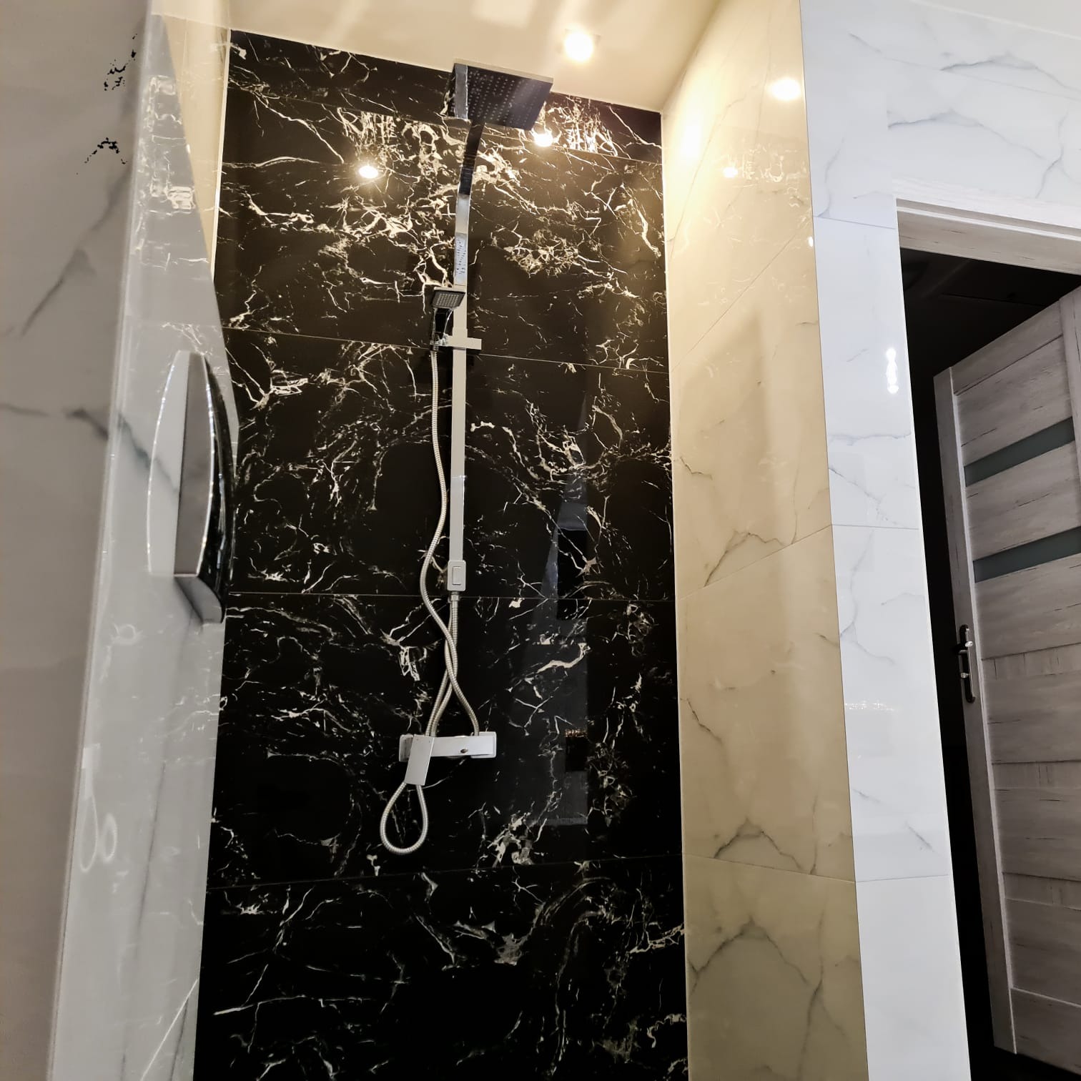 Nowoczesna kabina prysznicowa z czarnymi płytkami imitującymi marmur i chromowaną armaturą, obok jasne płytki i fragment drzwi.