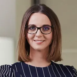 Portret uśmiechniętej kobiety w okularach i sukience w paski, z krótkimi brązowymi włosami, na rozmytym tle.