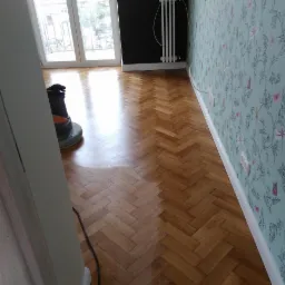 Parkiet ułożony w jodełkę, świeżo po zabiegu renowacji, z widocznym połyskiem i maszyną do obróbki podłóg w tle, na ścianie wzorzysta tapeta.