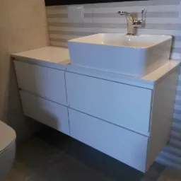 Biała, kwadratowa umywalka nablatowa z chromowaną baterią, osadzona na szafce z czterema szufladami o gładkich frontach, w łazience wykończonej płytkami w odcieniach beżu i szarości.