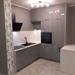 Szara, nowoczesna kuchnia z białymi płytkami na ścianie, czarnym okapem i blatem, oraz kolumną z tapetą w motywy drzewne.