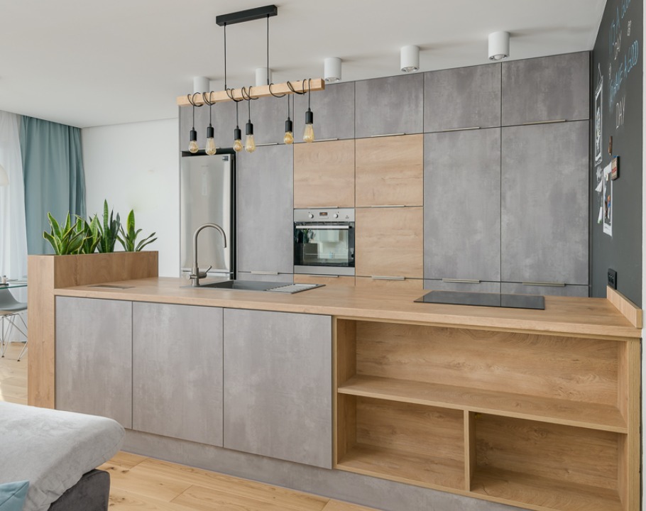 Nowoczesna kuchnia z szarymi frontami i drewnianym blatem, oświetlona wiszącymi lampami z żarówkami Edisona. Na blacie zlew i płyta indukcyjna, z boku półki.