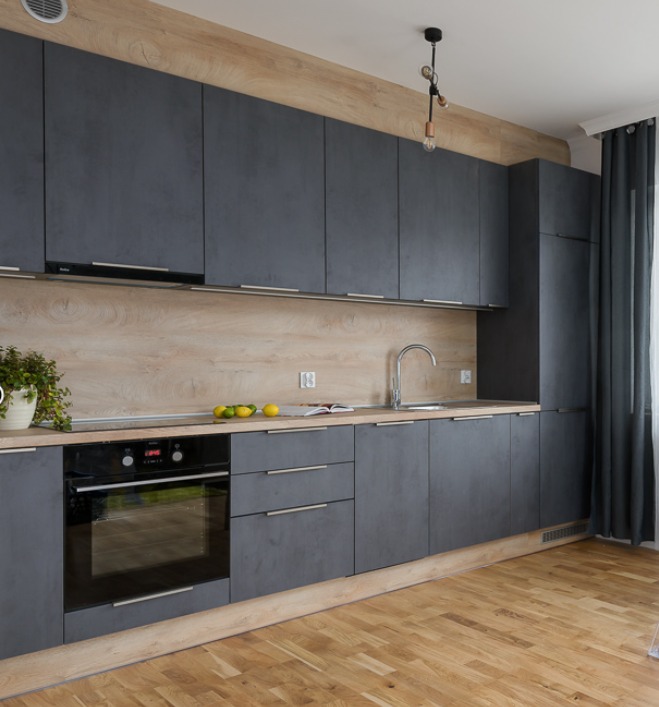 Nowoczesna kuchnia z szarymi matowymi frontami, drewnianym blatem i panelem ściennym. Widoczny piekarnik, zlew i oświetlenie. Podłoga z jasnego drewna.