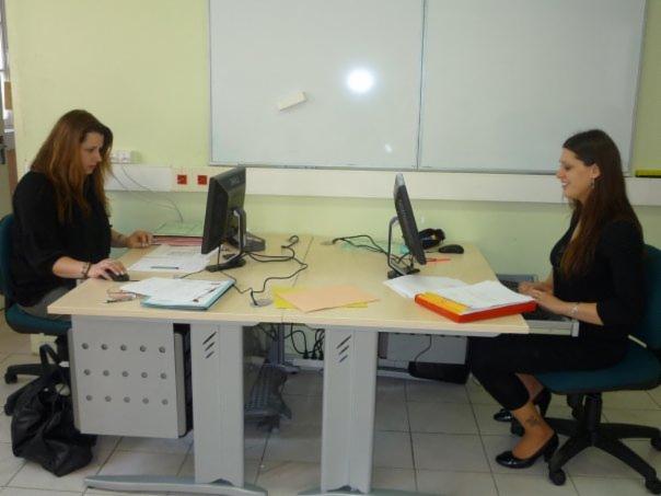 Dwie kobiety pracują przy biurkach z komputerami, przeglądając dokumenty i korzystając z monitorów, na tle białej tablicy.