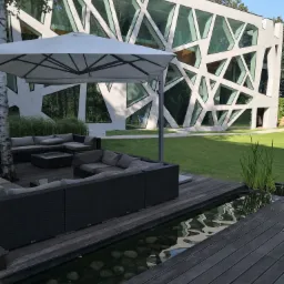 Nowoczesna elewacja budynku z geometrycznymi przeszkleniami w Warszawie, widoczna z tarasu z meblami ogrodowymi i oczkiem wodnym.