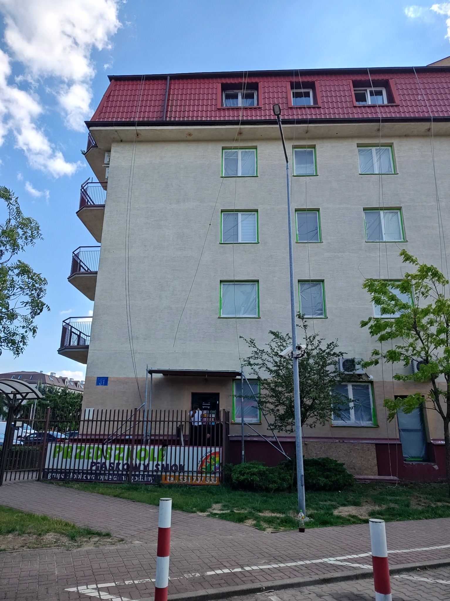 Przygotowanie elewacji budynku do malowania: okna zabezpieczone folią, widoczne liny do prac wysokościowych, fragment ogrodzenia z reklamą przedszkola.