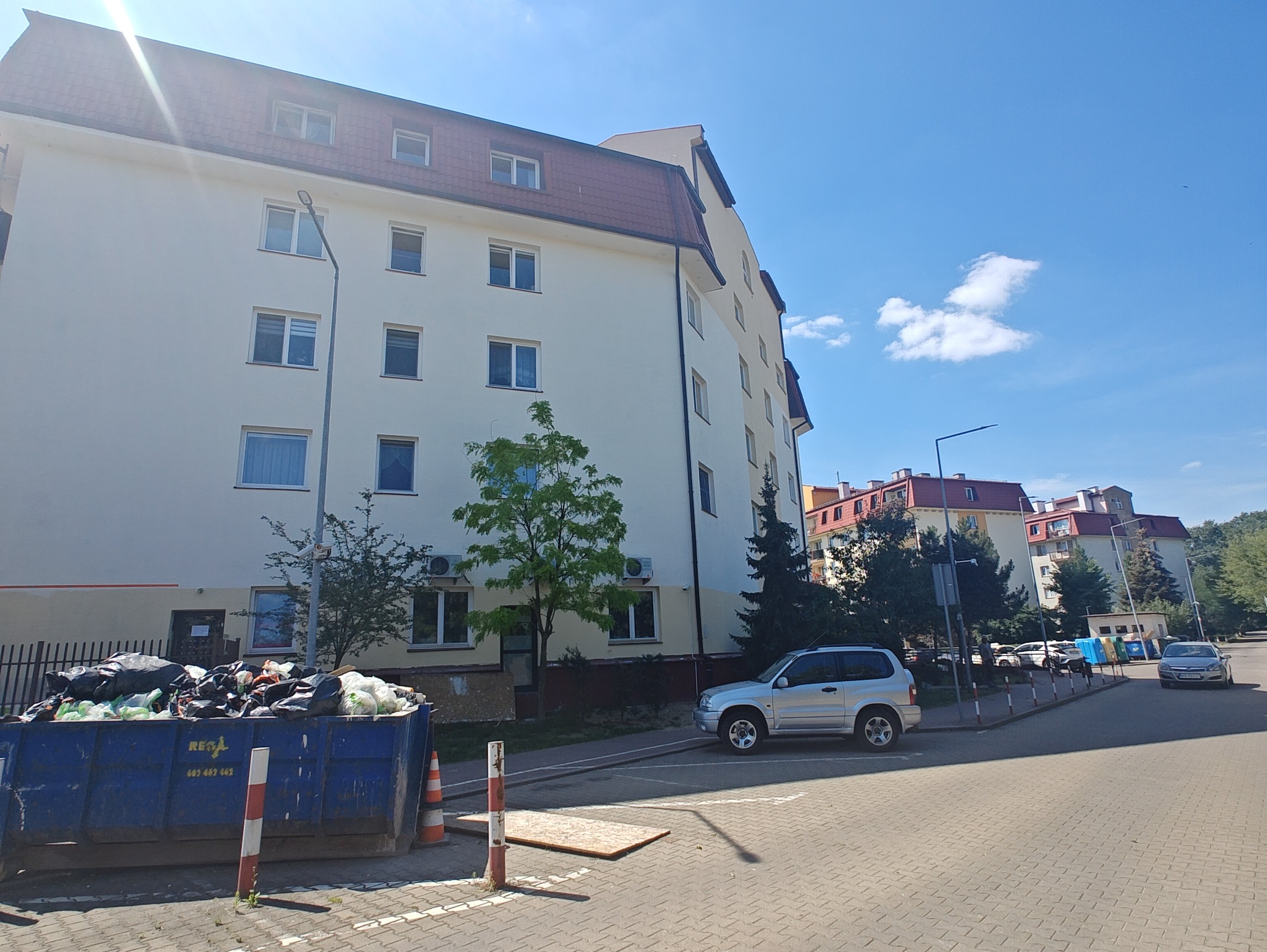 Wielopiętrowy budynek mieszkalny z jasną elewacją i czerwoną dachówką, widoczny z poziomu ulicy, z kontenerem na śmieci wypełnionym odpadami w pierwszym planie, srebrnym SUVem zaparkowanym obok...