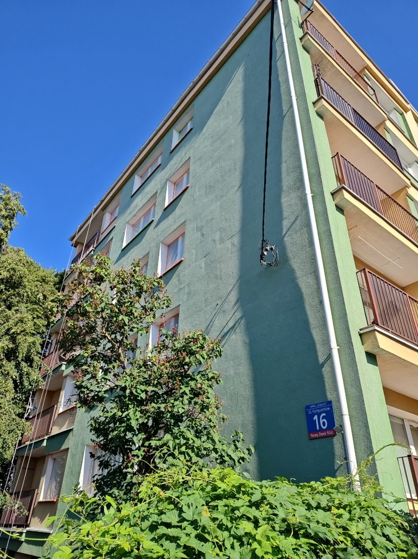 Wielopiętrowy blok mieszkalny z balkonami, pomalowany na zielono, z widoczną instalacją elektryczną biegnącą wzdłuż elewacji i tabliczką z numerem 16. Częściowo zasłonięty przez zieleń.