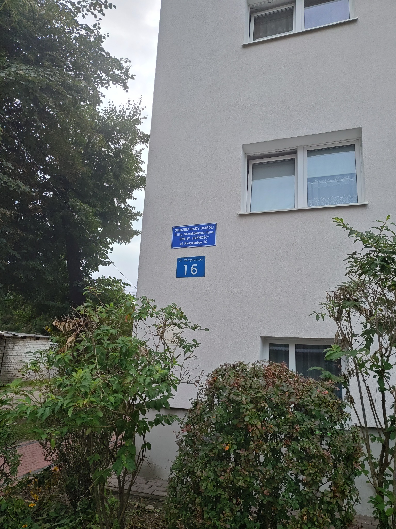 Biały budynek z tabliczką informacyjną 'Siedziba Rady Osiedli' i numerem 16 przy ulicy Partyzantów, częściowo zasłonięty przez krzewy i drzewa.