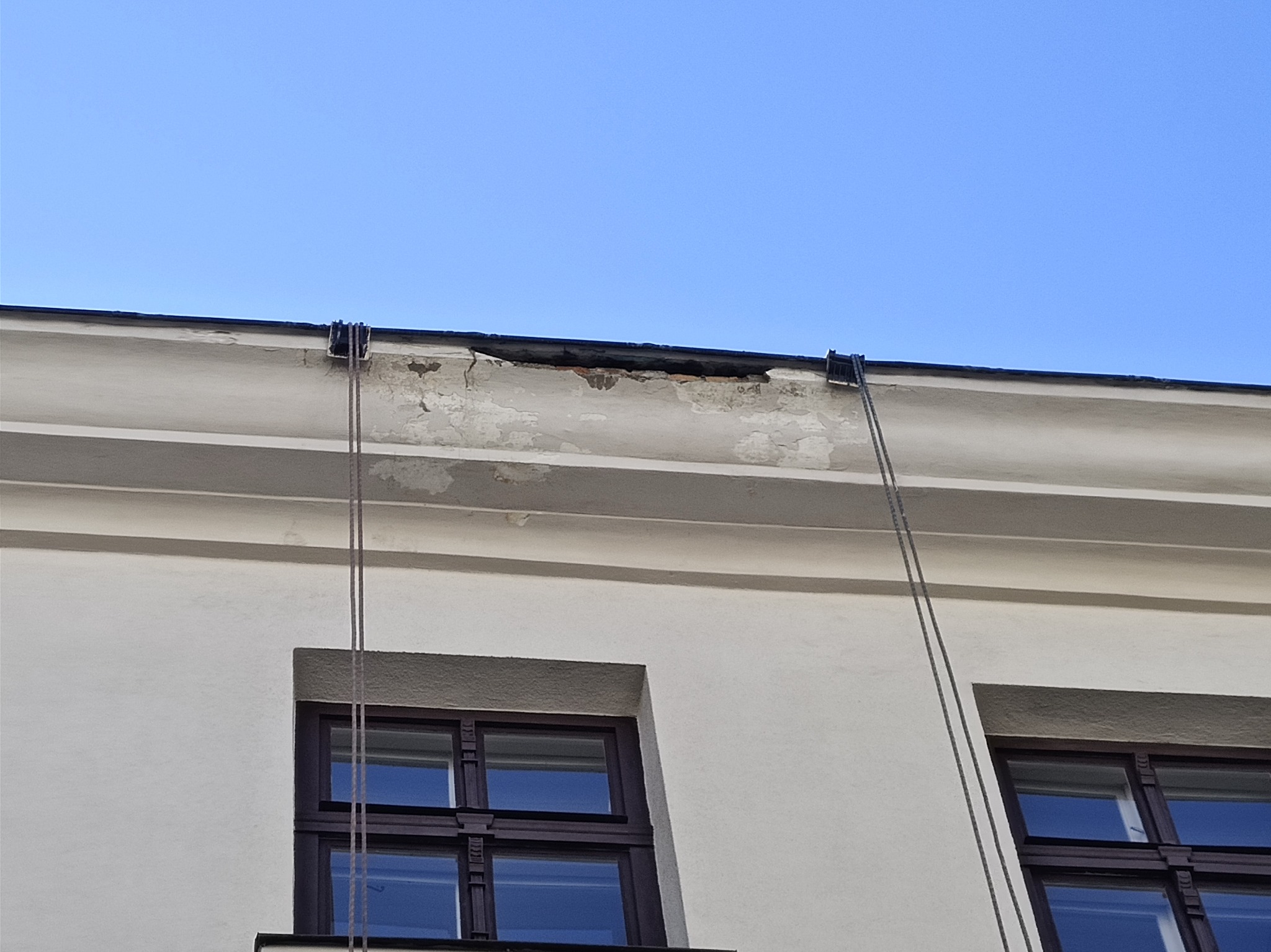 Fragment elewacji budynku z widocznymi uszkodzeniami tynku i fragmentem liny zabezpieczającej, na tle błękitnego nieba.