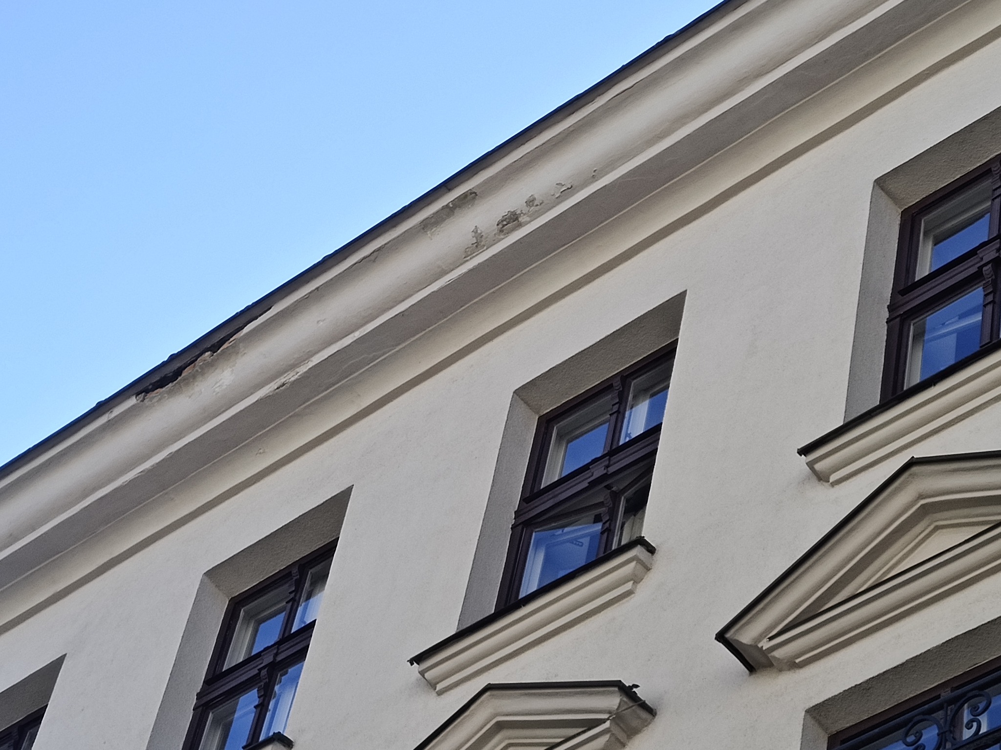 Fragment elewacji budynku z widocznymi oknami i uszkodzeniami tynku przy gzymsie, sfotografowany pod kątem, z otwartym oknem po prawej stronie.