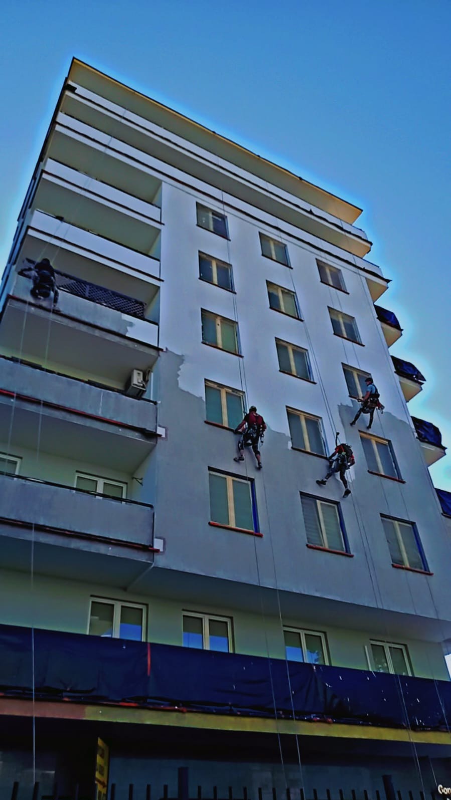 Malowanie elewacji bloku przez alpinistów. Częściowo pomalowana ściana, widoczne liny i zabezpieczenia. Ujęcie z dołu, błękitne niebo w tle.