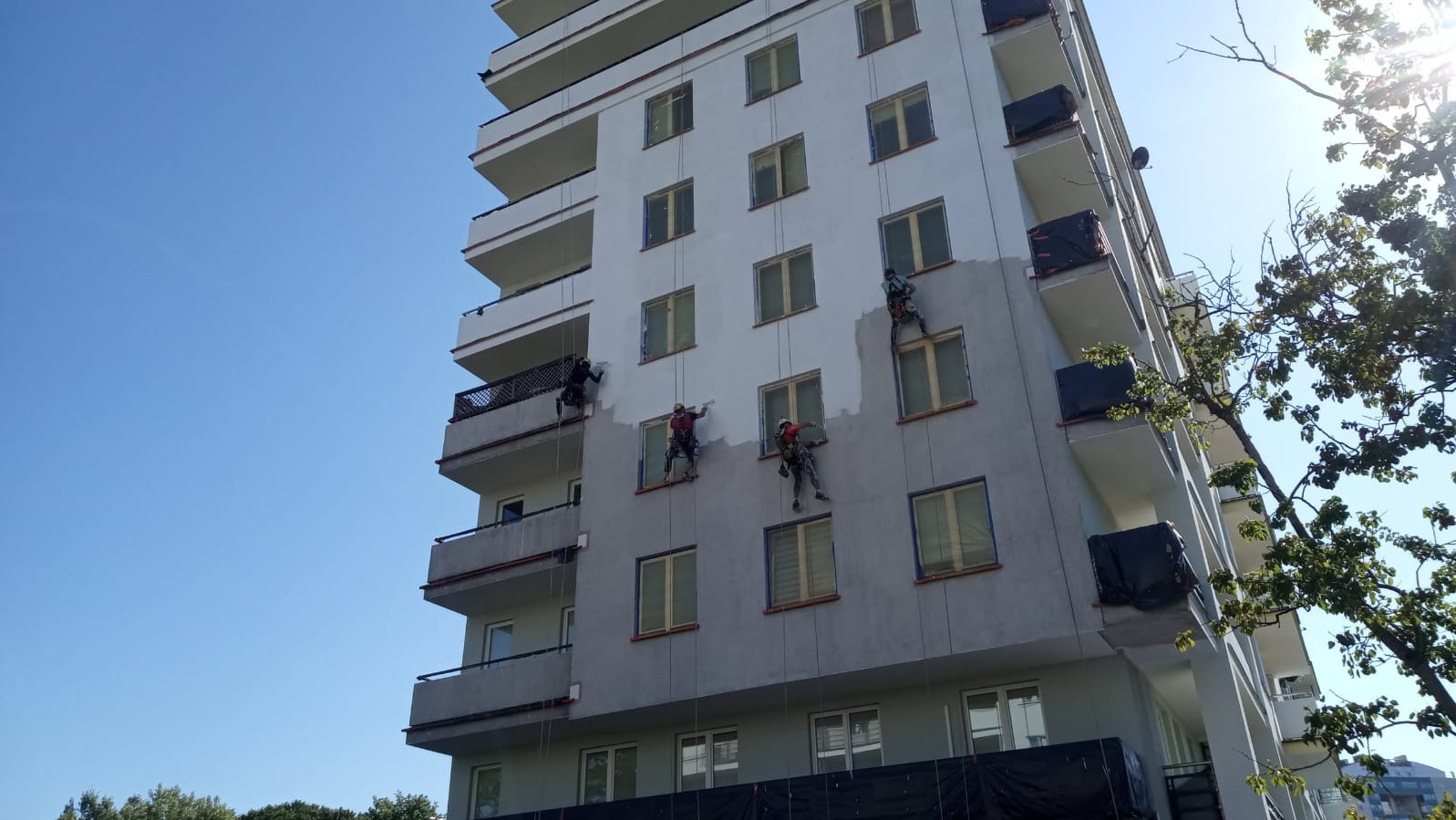 Malowanie elewacji bloku przez alpinistów na linach. Część budynku pomalowana na szaro, okna zabezpieczone. Błękitne niebo w tle, widok z dołu.