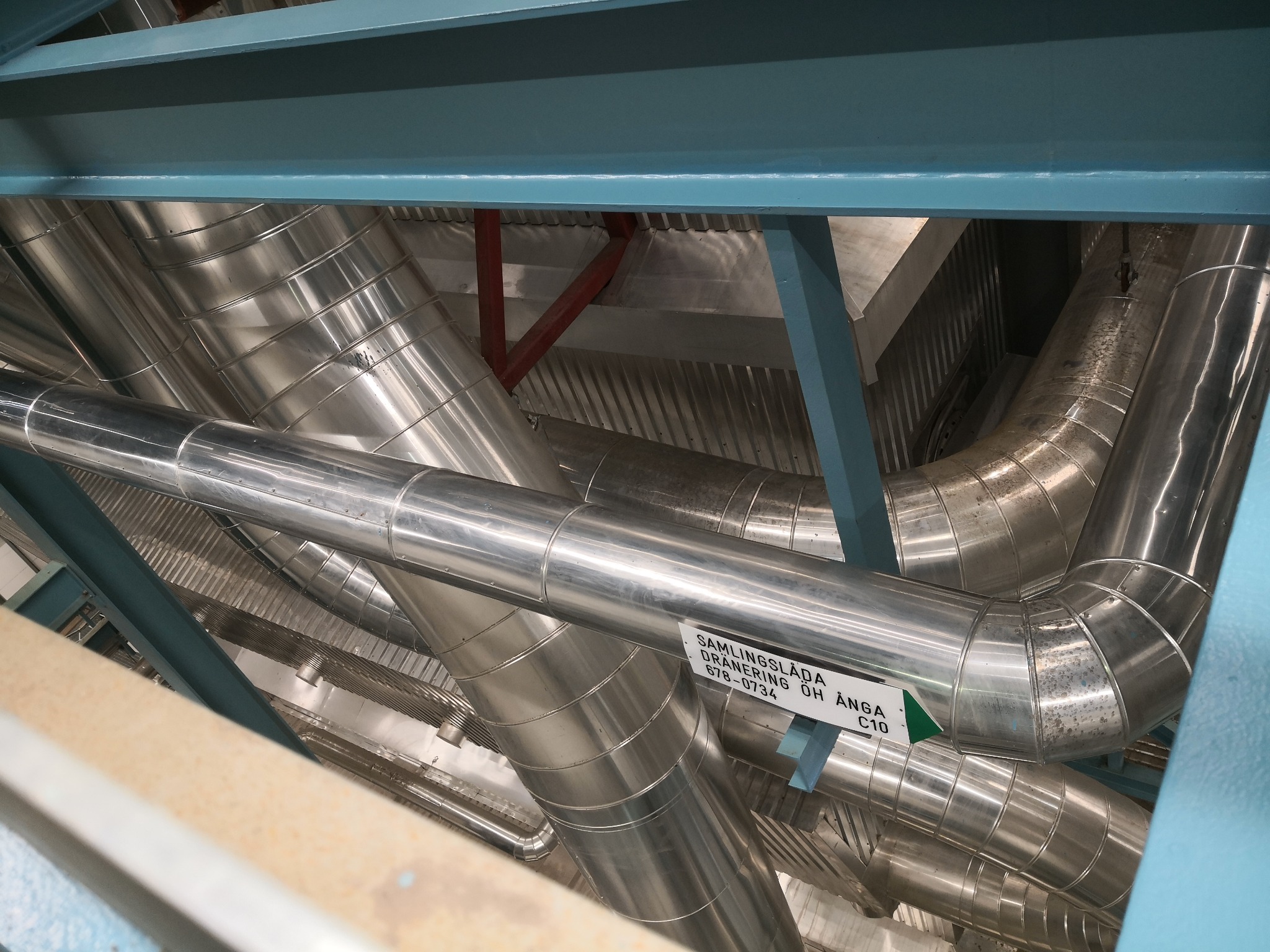 Widok z dołu na skomplikowany układ rur izolowanych termicznie, biegnących pod sufitem, z widoczną stalową konstrukcją nośną i tabliczką informacyjną z napisem 'SAMLINGSLÅDA DRANERING ÖH ÅNGA...
