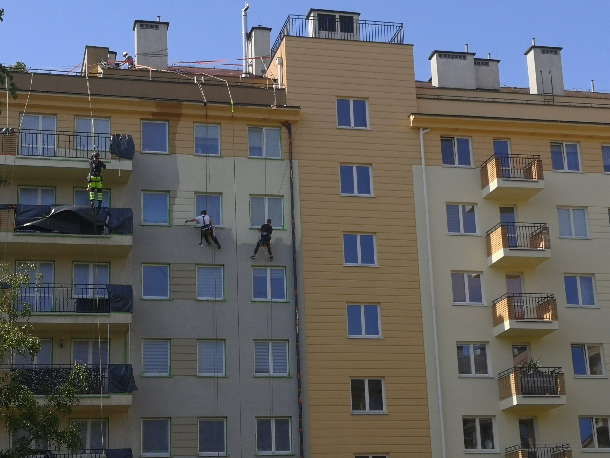 Trzech pracowników na linach zabezpieczających podczas prac na wysokościach, dwóch maluje elewację budynku, jeden kontroluje z góry, widoczne zabezpieczenia okien i balkonów folią.