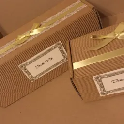 Dwa tekturowe pudełka z prążkowanej tektury, ozdobione złotymi wstążkami i etykietami z napisem 'Thank You'. Jedno pudełko ma dodatkowo koronkową tasiemkę.