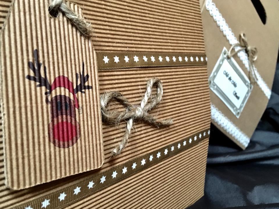 Brązowe pudełko z tektury falistej, ozdobione wstążką z gwiazdkami i sznurkiem jutowym, etykieta z reniferem w czapce Mikołaja, w tle torba prezentowa z koronką.